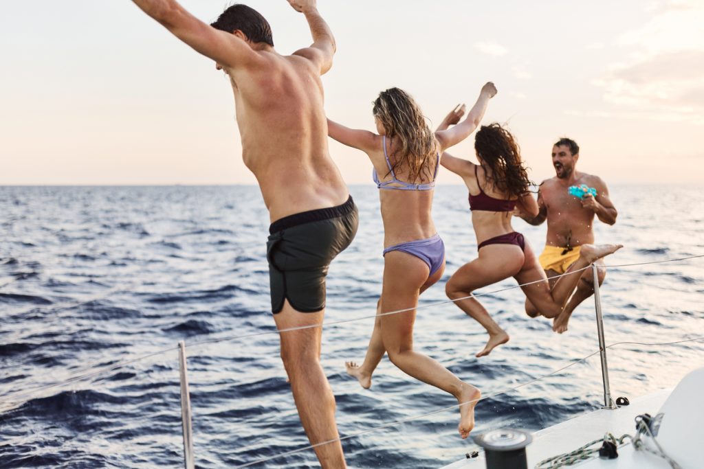 Back view of carefree friends having fun while jumping with raised arms from a boat in summer day. Copy space.
